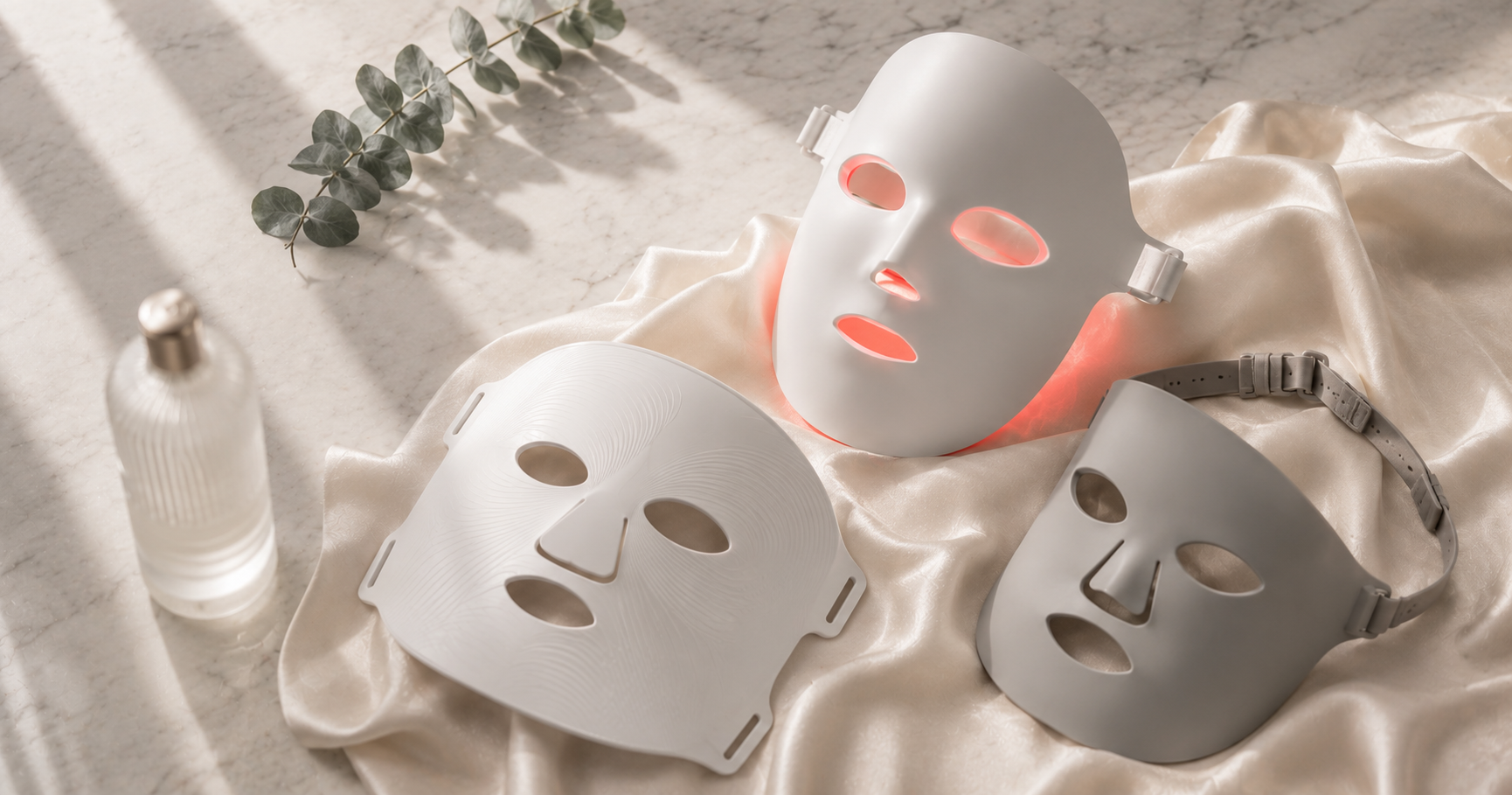 Several luxury LED face masks including a red-glowing rigid mask and flexible silicone masks arranged on Calacatta marble with ivory silk